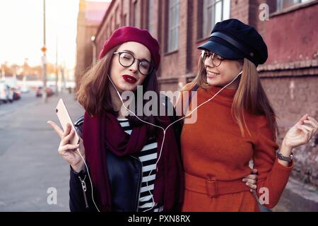 Two joyful young girls dancing while listening to music on smartphone, city outdoor. Stock Photo