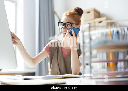 Young woman sitting at office table and pointing at computer screen while talking on the phone Stock Photo