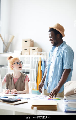 Young businesswoman sitting at her workplace and discussing something with her smiling colleague at atelier Stock Photo