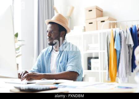 Young African stylish man sitting at the table and typing on keyboard and looking at computer screen at office Stock Photo
