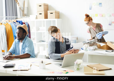 Young two men sitting at the table typing on computer and talking on the mobile phone with woman packing clothes in the background Stock Photo