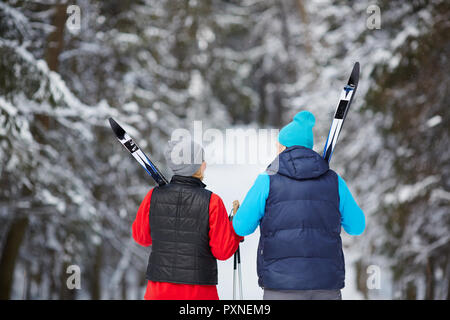 Back view of mature sporty couple in activewear carrying skiing equipment while moving in winter woods Stock Photo