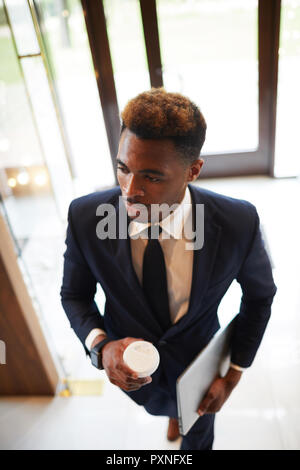 Young African businessman in suit holding coffee and laptop walking into an office building Stock Photo