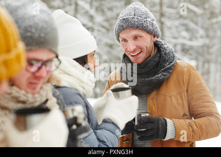Cheerful young friends in warm coats talking to each other and drinking tea from thermos mug in winter hike Stock Photo