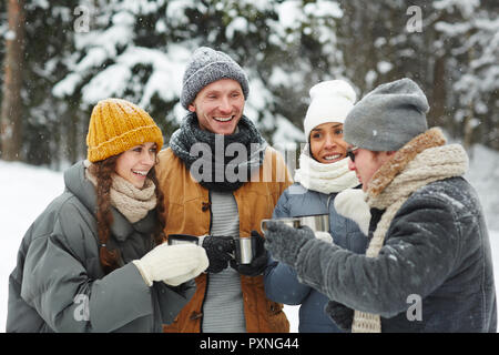 Happy excited young travelers in winterwear standing in forest and drinking hot tea while chatting at winter hike Stock Photo