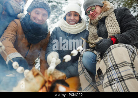Cheerful excited young multi-ethnic friends sitting together and roasting marshmallows on stick at camping, they looking at camera Stock Photo