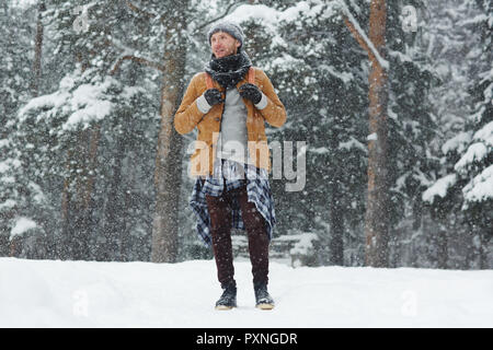 Content handsome young traveler in warm clothing standing on snow and looking around while contemplating nature and walking in forest Stock Photo