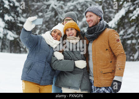 Group of cheerful excited young friends in puffy jackets laughing while taking selfie on smartphone in winter forest Stock Photo