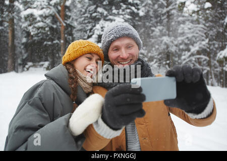 Jolly excited beautiful young couple in warm clothing standing in winter forest and smiling while taking selfie ion smartphone Stock Photo