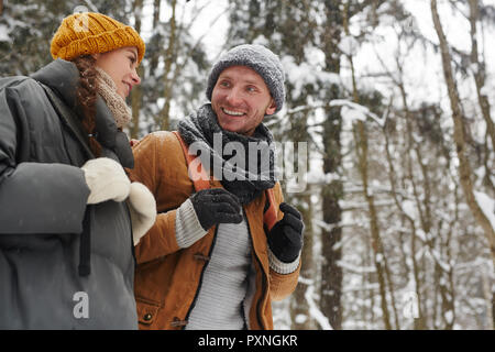 Cheerful positive young friends in warm hats wearing backpacks walking over forest and talking while hiking together in winter forest Stock Photo