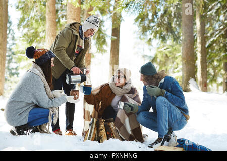 Full length portrait of four young  friends camping in winter forest sitting in circle round fire and pouring hot drinks Stock Photo