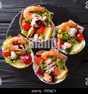Beautiful seafood cocktail salad of shrimp, baby octopus, mussels, squids and scallops with vegetables in sea shells close-up on a black table. top vi Stock Photo
