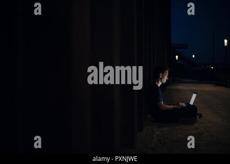 Mid adult man sitting cross-legged on ground, using laptop at night Stock Photo