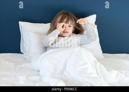 Little girl stretching in white bed, yawning and rubbing her eyes, awaking early in the morning Stock Photo
