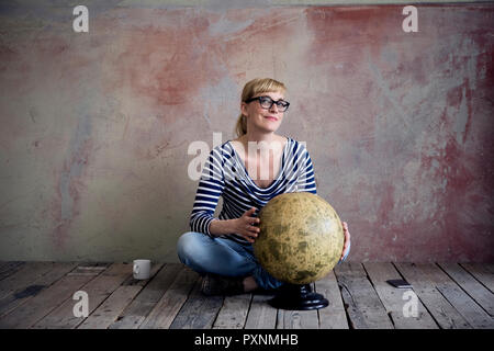 Smiling woman sitting on wooden floor in an unrenovated room with an old globe Stock Photo