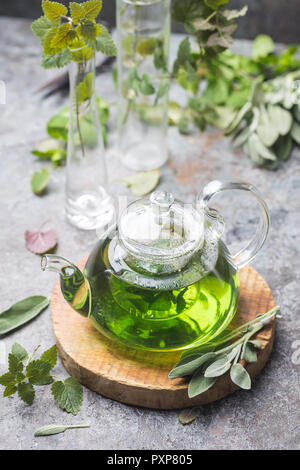 Hot herbal sedative mint tea drink in a glass teapot on wooden tray with fresh garden mint and sage leaves over gray background Stock Photo
