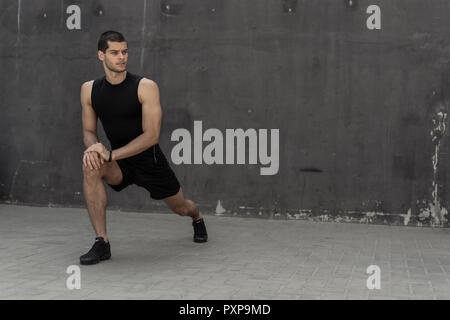 Handsome, athletic man warming up stretching before the training, with gray walls in the background. Healthy lifestyle and sportswear advertising. Stock Photo
