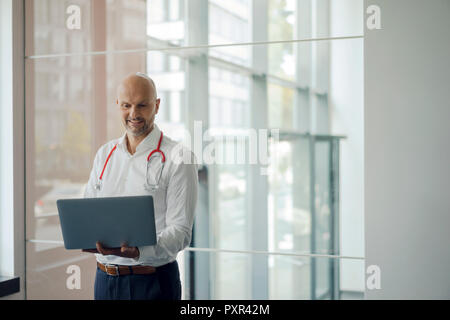 Doctor standing in hospital with stethoscope around his neck, holding laptop Stock Photo
