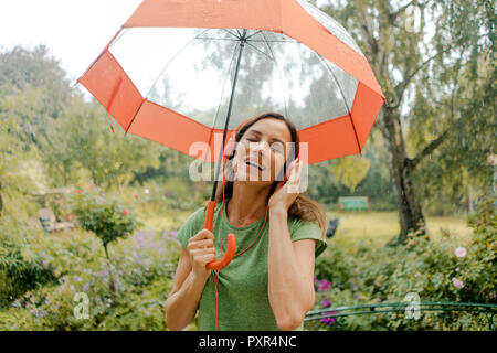 Happy mature woman standing in rain under umbrella listening to music with headphones Stock Photo