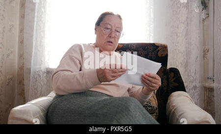 An elderly woman sitting in a chair and reading something, looking surprised not in a good way. Stock Photo