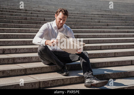 Smiling businessman sitting on stairs reading newspaper Stock Photo