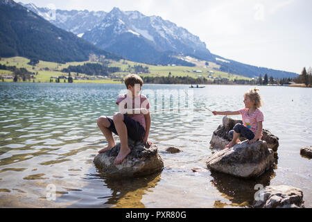 Austria, Tyrol, Walchsee, brother and sister sitting on boulders in the lake playing with sticks Stock Photo