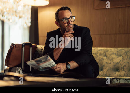 Portrait of thoughtful mature businessman sitting in hotel lobby with his luggage and looking away. Mature CEO waiting in hotel lobby holding newspape Stock Photo
