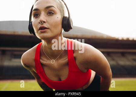 fitness woman training in a stadium bending forward with hands on knees. Female athlete doing workout wearing wireless headphones. Stock Photo