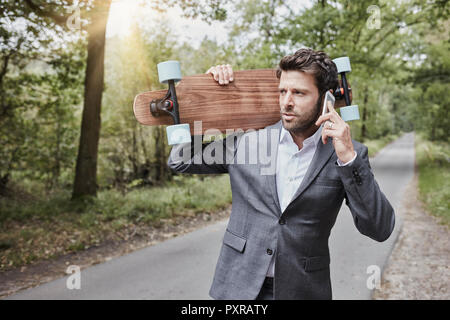 Businessman with skateboard talking on smartphone on rural road Stock Photo
