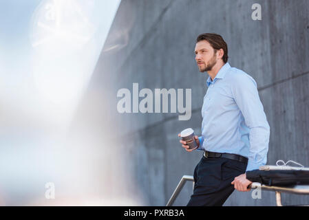 Businessman with laptop bag and takeaway coffee leaning against a railing Stock Photo