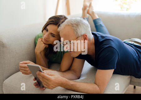 Smiling mature couple lying on couch at home sharing tablet Stock Photo