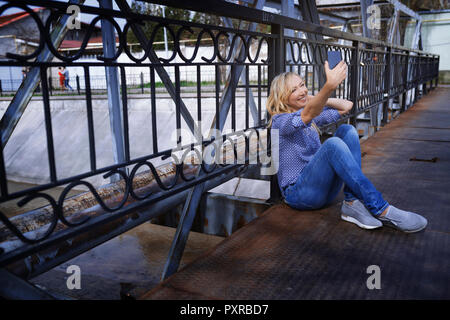 Blond woman sitting on bridge, taking selfie Stock Photo