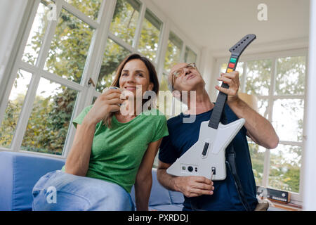 Mature couple sitting on couch at home with man playing toy electric guitar Stock Photo