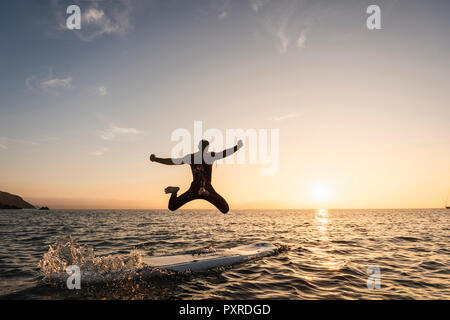Young man jumping from paddleboard into water at sunset Stock Photo