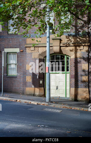 A white door into a brick and sandstone building on a sloping street. In front is a lamp post and a tree which are casting shadows in the brick wall. Stock Photo