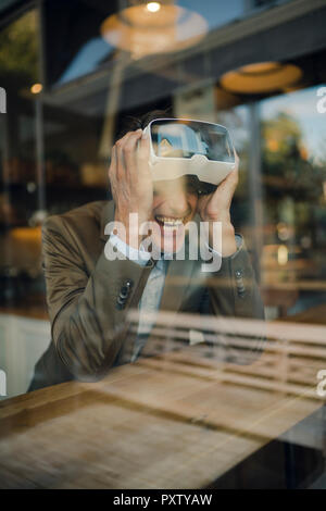 Mature businessman sitting in coffee shop, looking through VR glasses, laughing Stock Photo