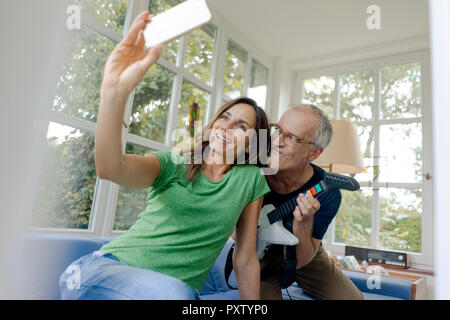Happy mature couple taking a selfie at home with man playing toy electric guitar Stock Photo