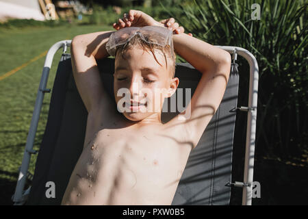 Wet boy wearing safety goggles lying on sun lounger in garden Stock Photo