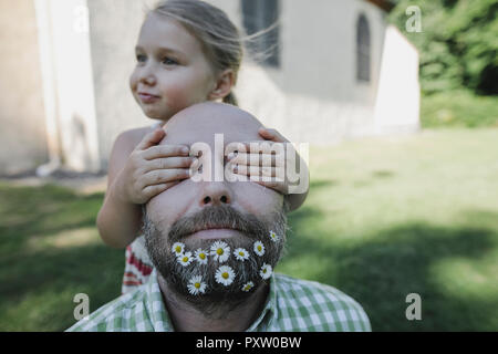 Little girl's hands covering eyes of mature man with daisies in his beard Stock Photo