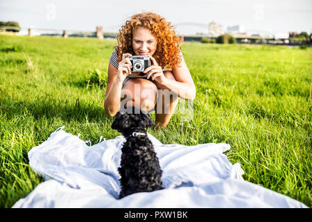 Germany, Cologne, portrait of smiling young woman on meadow taking picture of her dog Stock Photo