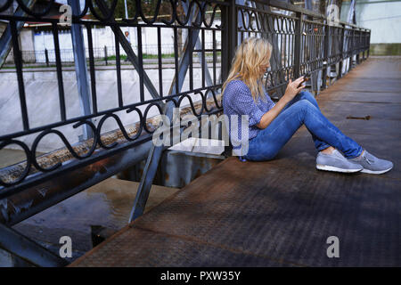 Blond woman sitting on bridge, using smartphone Stock Photo