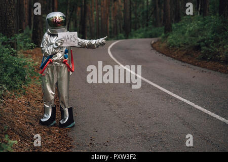 Spaceman hitchhiking to Mars, standing on road in forest Stock Photo