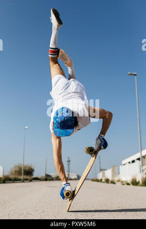 Back view of man in stylish sportive outfit standing on skateboard upside down against blue sky Stock Photo