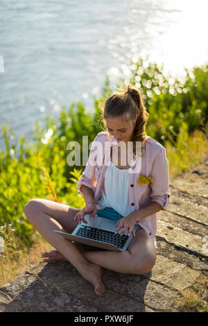 Woman sitting at the riverside in summer using laptop Stock Photo