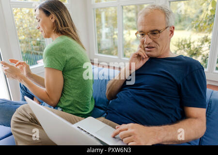 Mature couple sitting on couch at home with man using laptop and woman using cell phone Stock Photo