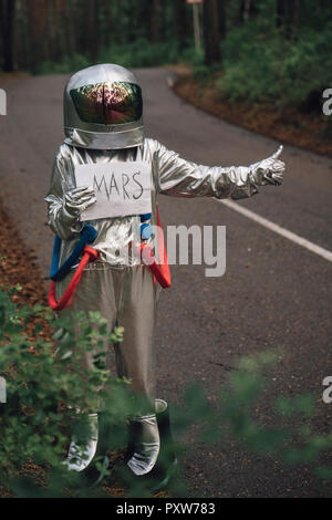 Spaceman hitchhiking to Mars, standing on road in forest Stock Photo