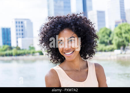 Germany, Frankfurt, portrait of smiling young woman with curly hair in front of Main River Stock Photo