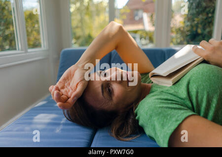 Smiling mature woman lying on couch at home with book Stock Photo