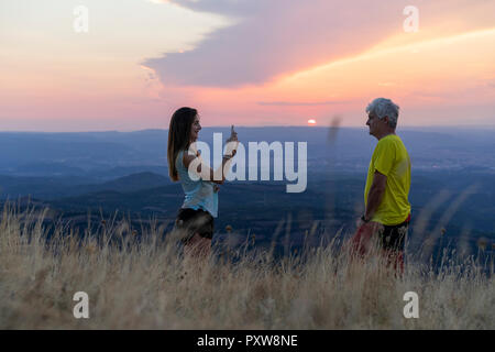 Spain, Catalonia, Montcau, senior father and adult daughter taking a cell phone picture on top of hill during sunset Stock Photo