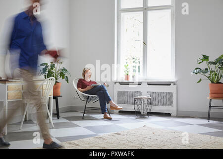 Mature woman reading magazine at home with man passing by Stock Photo